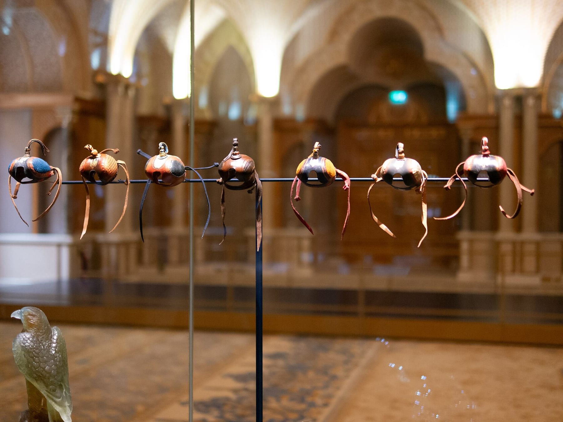 Seven leather falconry hoods on a display rail inside a vaulted hall at Qasr Al Watan, with a glass falcon figure at lower left, part of the palace visit priced on this page