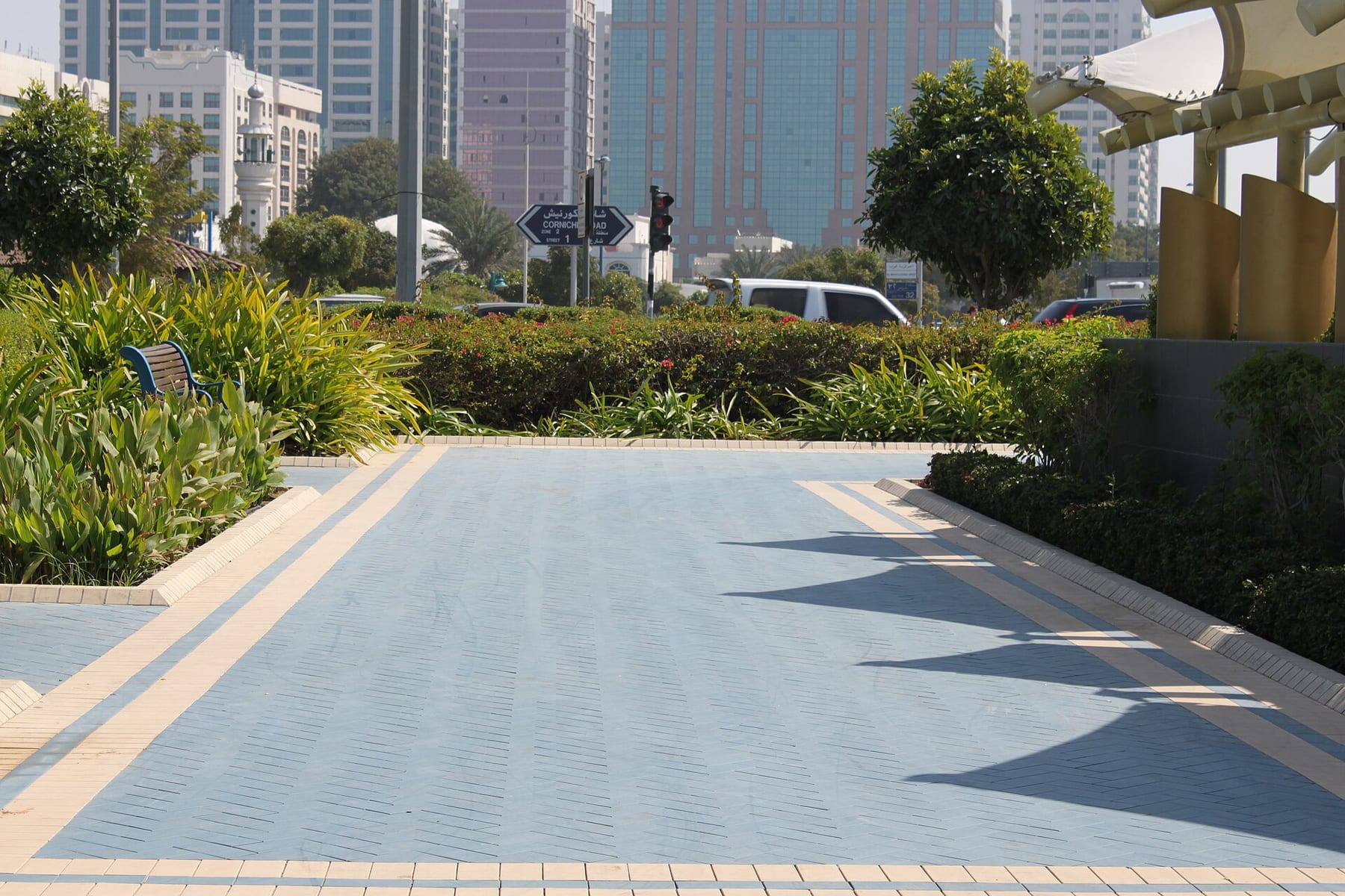 A blue-paved pedestrian path on the Corniche in Abu Dhabi, with garden beds and city-centre towers behind