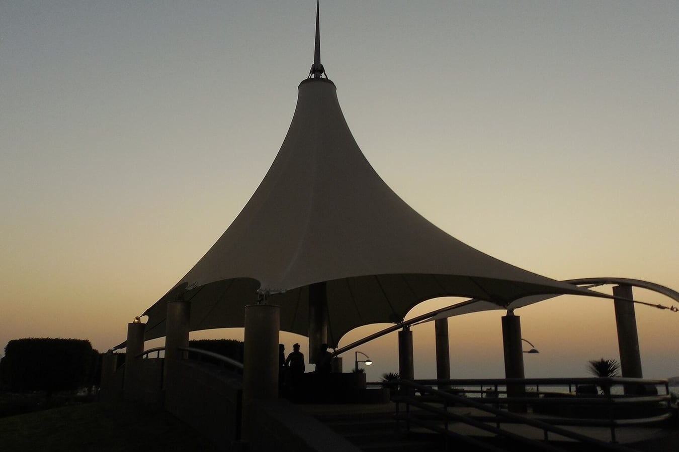 A white tensile shade roof on the Abu Dhabi Corniche in silhouette against the sky at dusk, with people standing beneath it