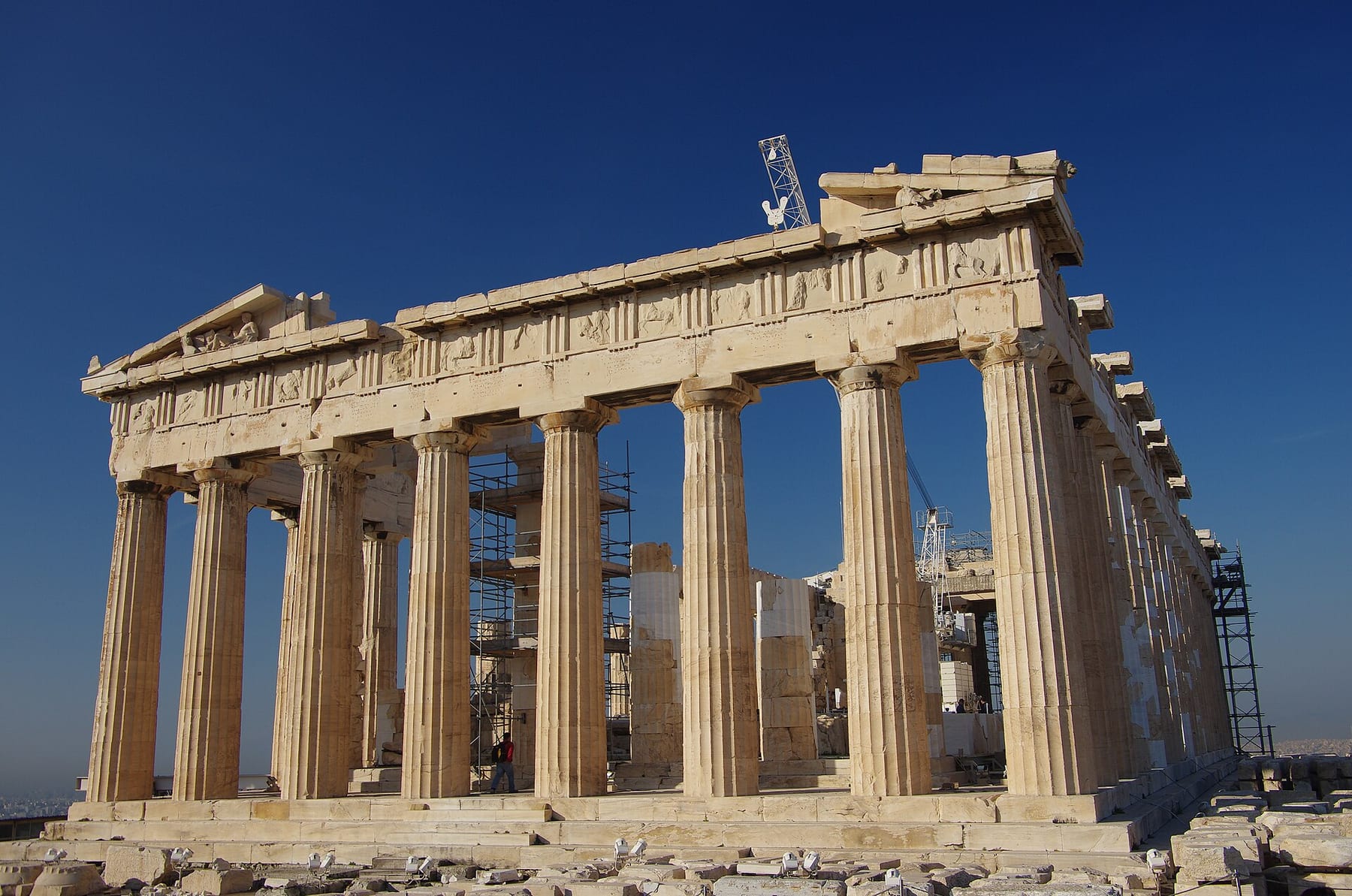 The Parthenon seen from the north-east on the Acropolis, its Doric columns and carved frieze against a deep blue sky, with restoration scaffolding and a crane tip at the rear