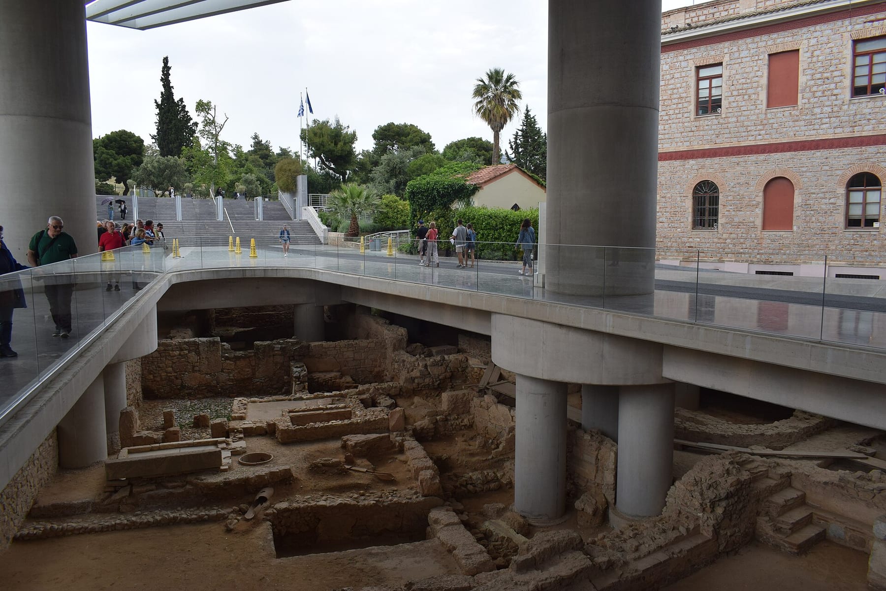 The glass-floored walkway outside the Acropolis Museum entrance, with excavated ancient foundations visible below and visitors walking under the museum’s concrete pillars