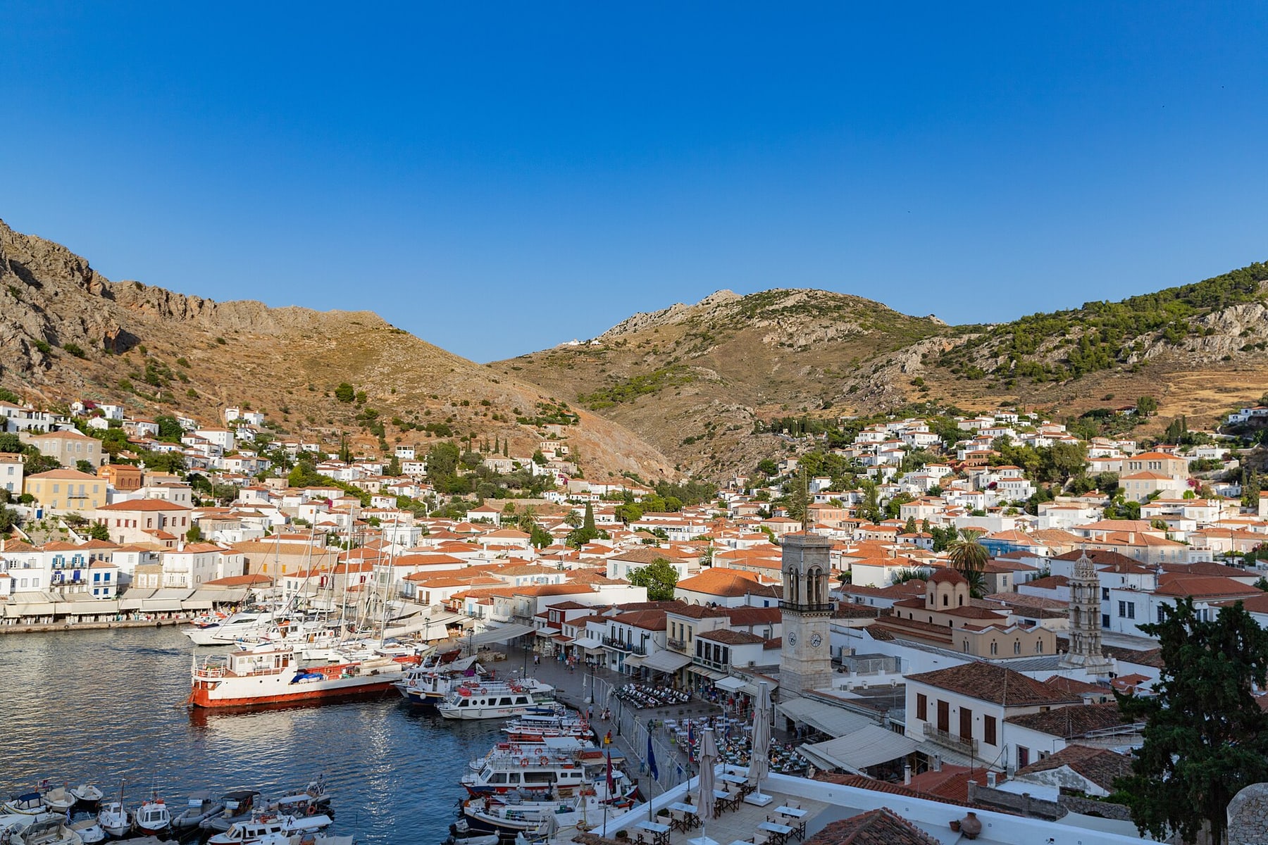 Hydra’s harbour with moored boats below a clock tower, and white and terracotta houses climbing the hillside
