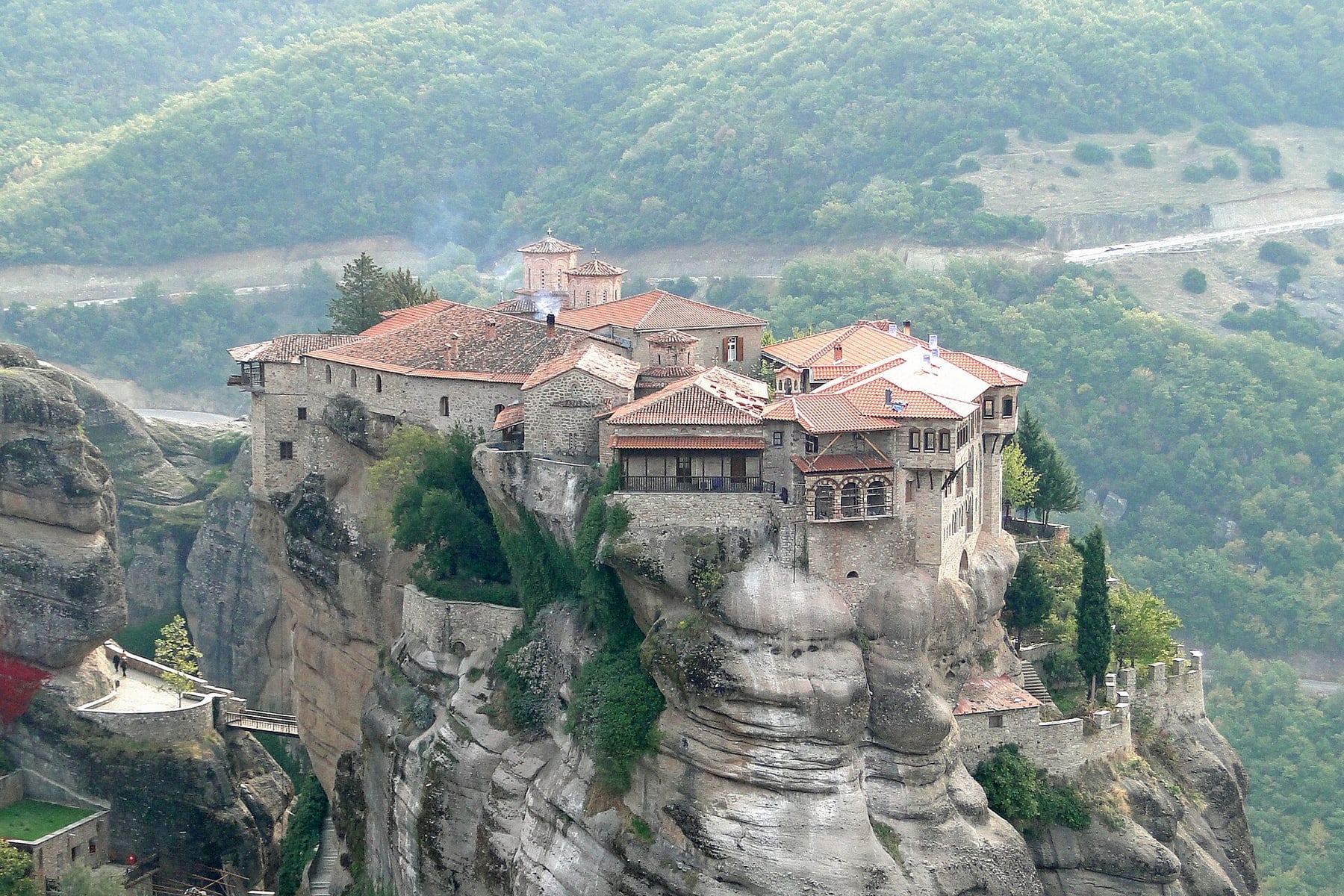 Varlaam Monastery on a tall rock pillar at Meteora, its red-tiled roofs and stone walls above forested hills