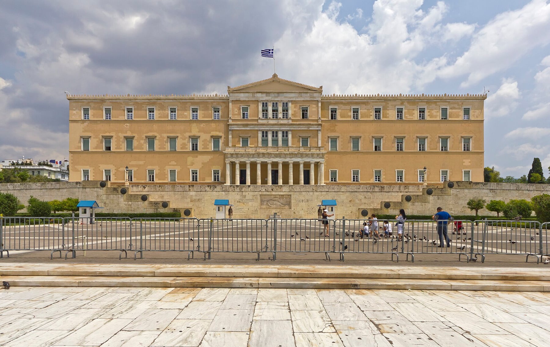 The Hellenic Parliament in Athens, a long neoclassical building behind the Tomb of the Unknown Soldier, with the Greek flag flying above Syntagma Square