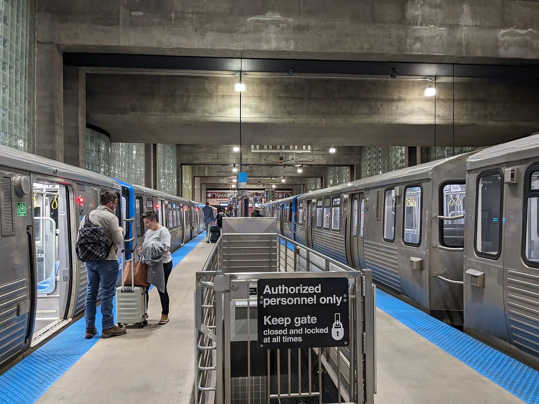Passengers with luggage on the platform beside a CTA Blue Line train at O’Hare station, with a second train on the far track