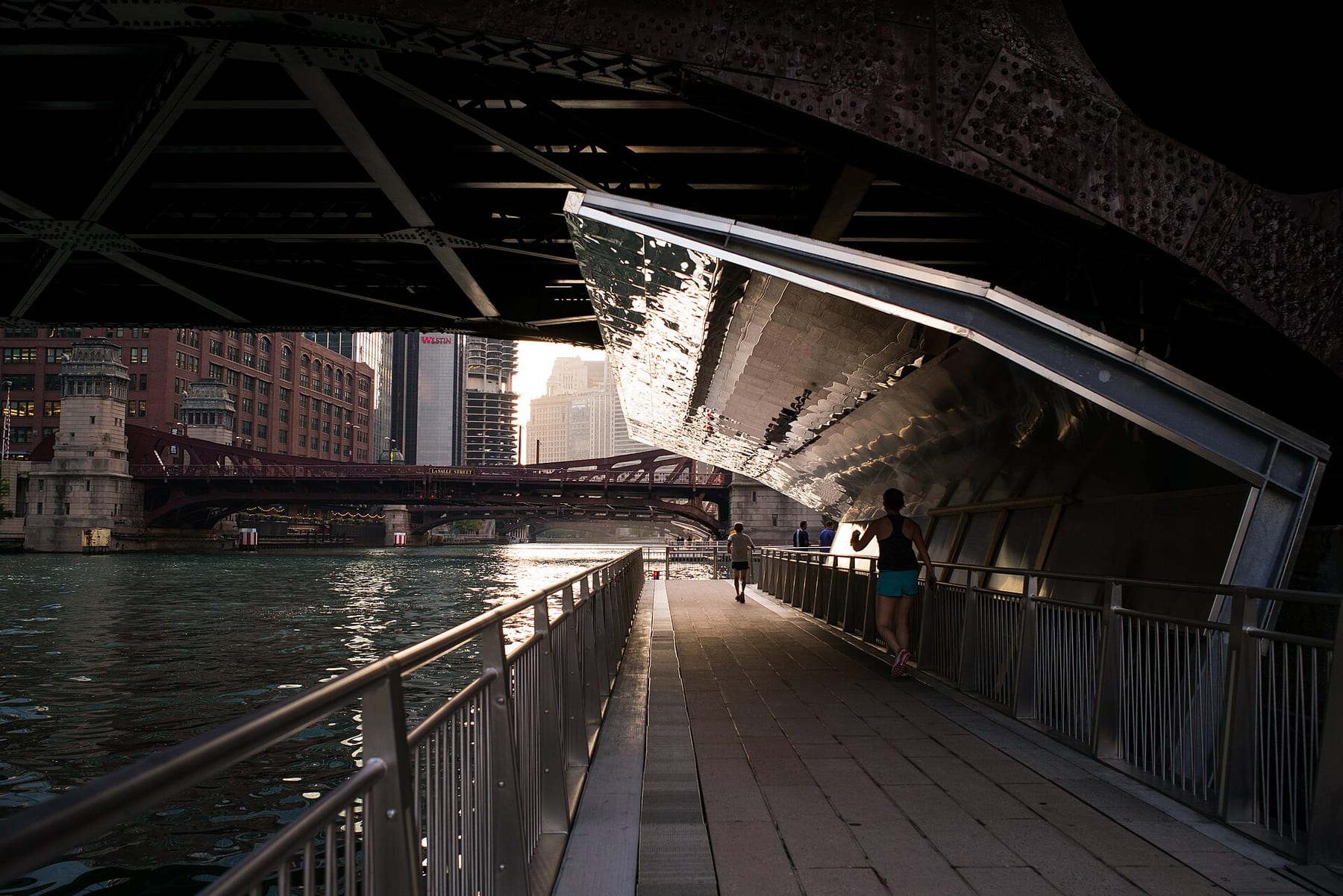 The Chicago Riverwalk passing under a bridge in low sun, with a runner on the path beside the river
