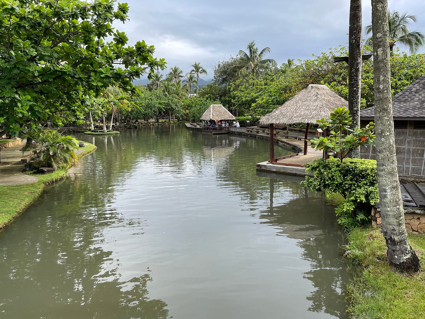 A lagoon channel at the Polynesian Cultural Center in Lāʻie, lined with palms and thatched island houses under a cloudy sky, the first ticket in the booking order