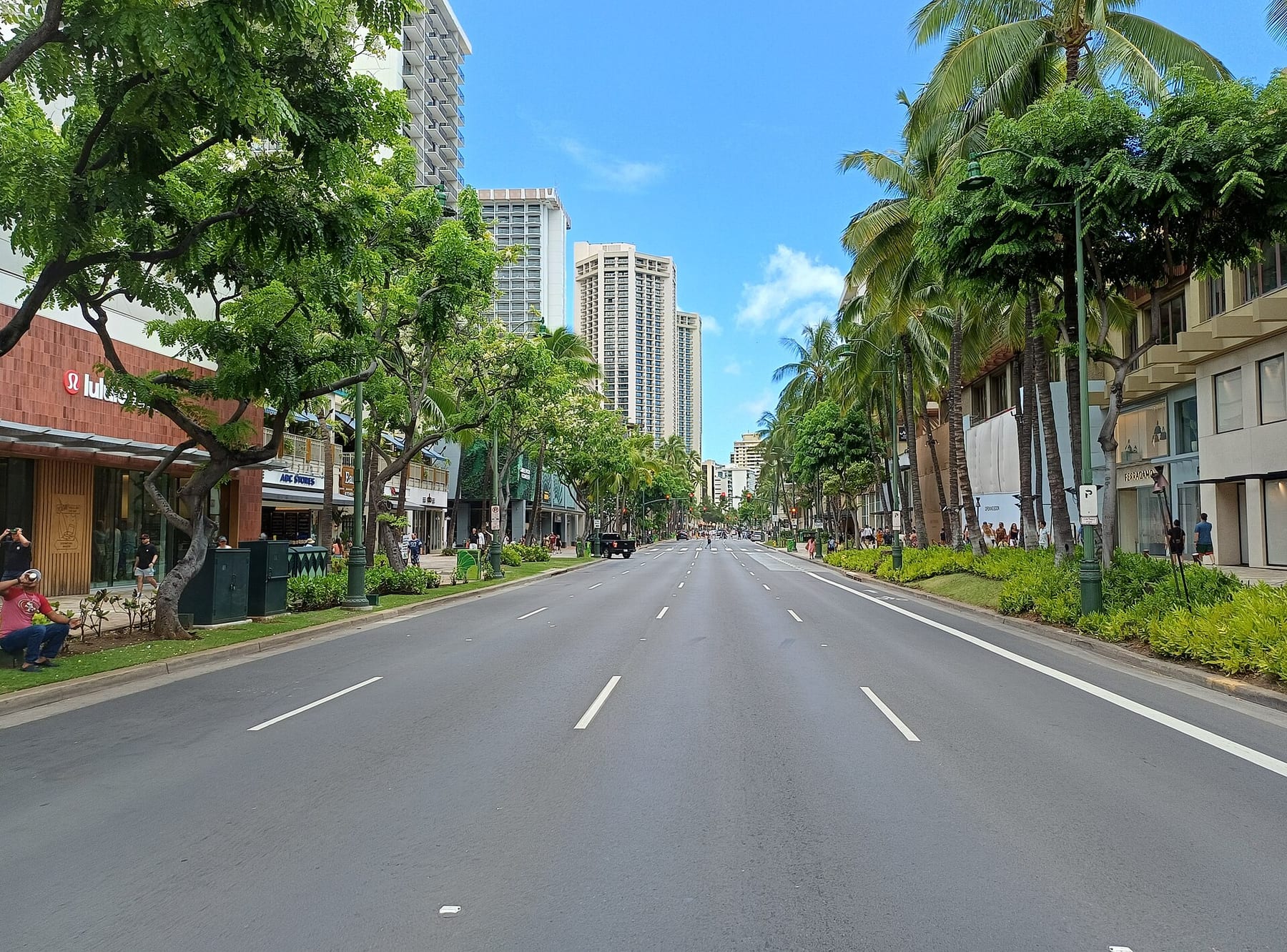 Kalakaua Avenue in Waikiki on a clear day, a wide empty road lined with palm and shade trees and high-rise buildings in the distance