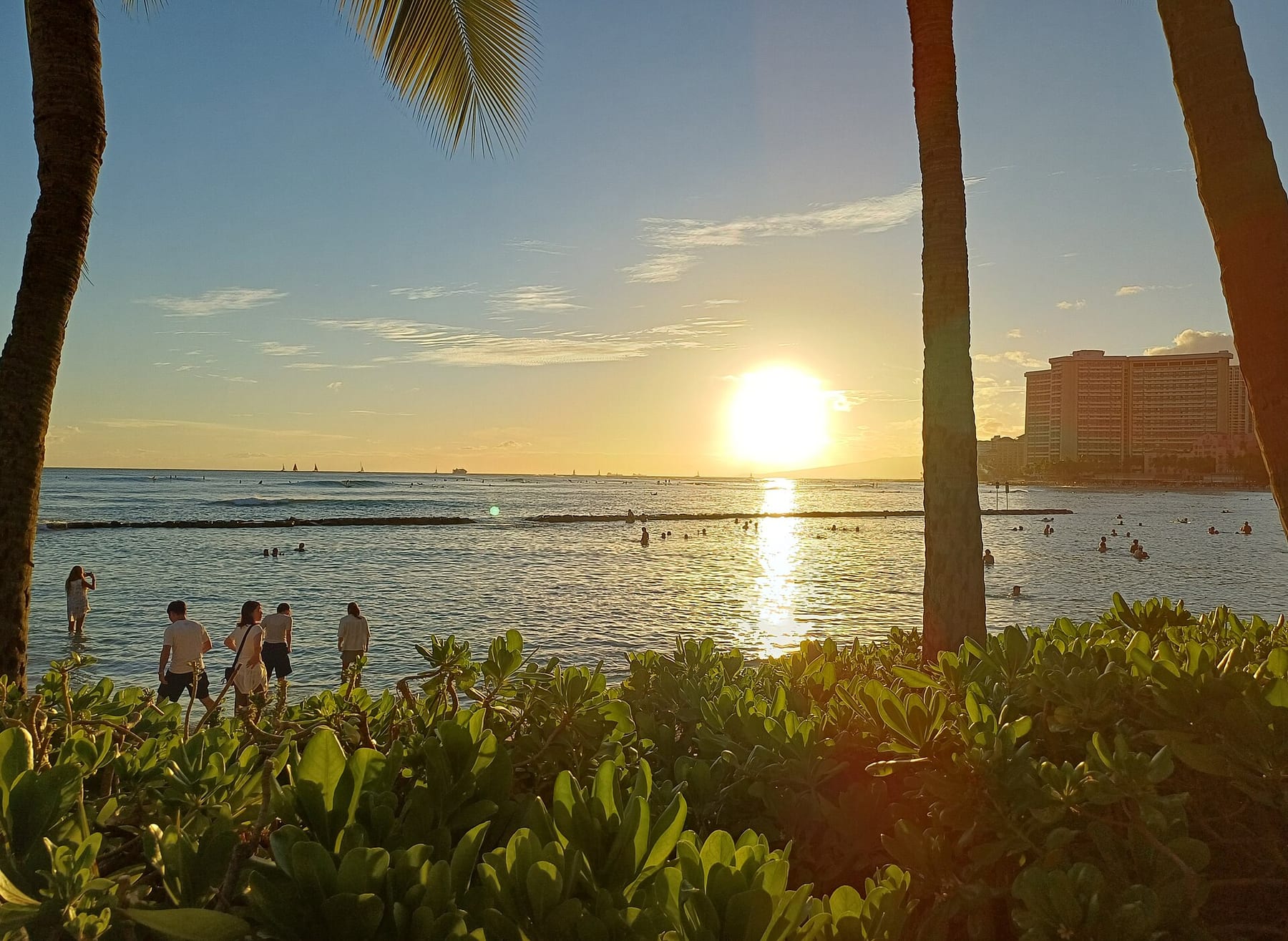 The sun low over the water at Kuhio Beach, with palm trunks in the foreground, a breakwater across the bay and swimmers in the shallows