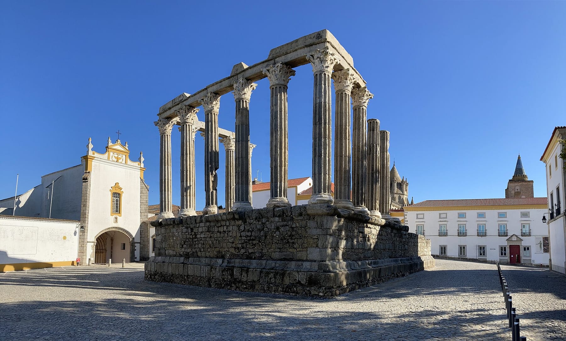 The Roman Temple of Évora, its Corinthian columns on a stone platform beside a white chapel under a blue sky
